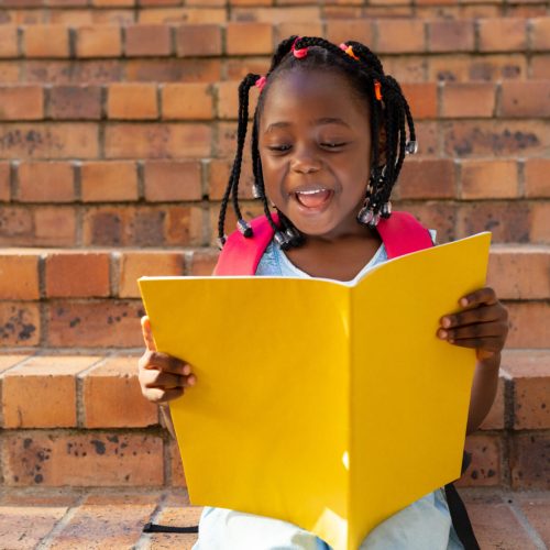 Happy african american schoolgirl sitting on stairs and reading book at elementary school. Education, childhood, development, learning and school, unaltered.