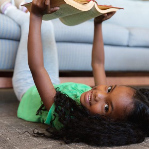Portrait of african american elementary girl reading book while lying on floor with legs over couch. unaltered, education, childhood, relaxation, reading, studying and school concept.